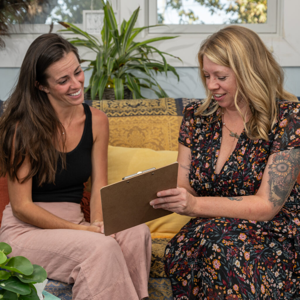 Two women sitting together on a couch, smiling and looking down at a wooden clipboard held by the woman on the right, who wears a floral dress and has tattooed arms. The woman on the left has long brown hair, wears a black tank top and pink pants. Behind them is a cozy room with a plant, patterned yellow throw pillows, and a window with greenery visible outside.
