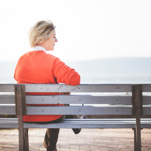 Woman with short blonde hair sitting alone on a bench, viewed from behind, wearing an orange cardigan over a white top and dark pants. She's turned slightly to the side, gazing out at a hazy waterfront or coastal view, with a soft, contemplative expression. The sky and water blend into a pale, misty backdrop, and a paved walkway with buildings faintly visible stretches along the horizon.